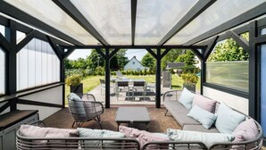 A covered patio area with a sofa, chairs, and a table, overlooking a garden and a house.