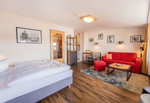 Une chambre d’hôtel moderne avec un lit, un canapé rouge, une petite table et un bureau. Les murs sont ornés de tableaux encadrés et le sol est en bois.