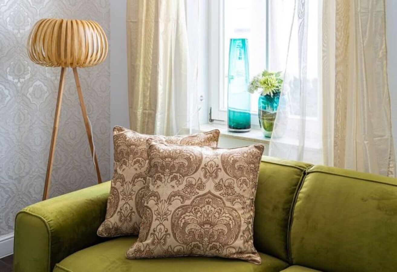 A green sofa with patterned cushions, a wooden floor lamp, and a window with sheer curtains and a potted plant.