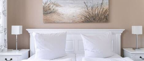 A neatly made bed with white bedding and two pillows, flanked by white nightstands with lamps, in a room with a beige wall and a beach-themed painting above the bed.