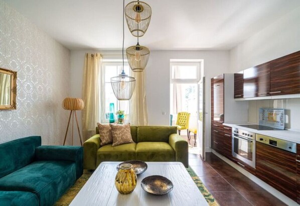 A modern living room with a green sofa, a white coffee table with decorative items, and a kitchen with wooden cabinets and a built-in oven. - Villa Meeresgold `Strandidyll` Premium Suite (Binz)