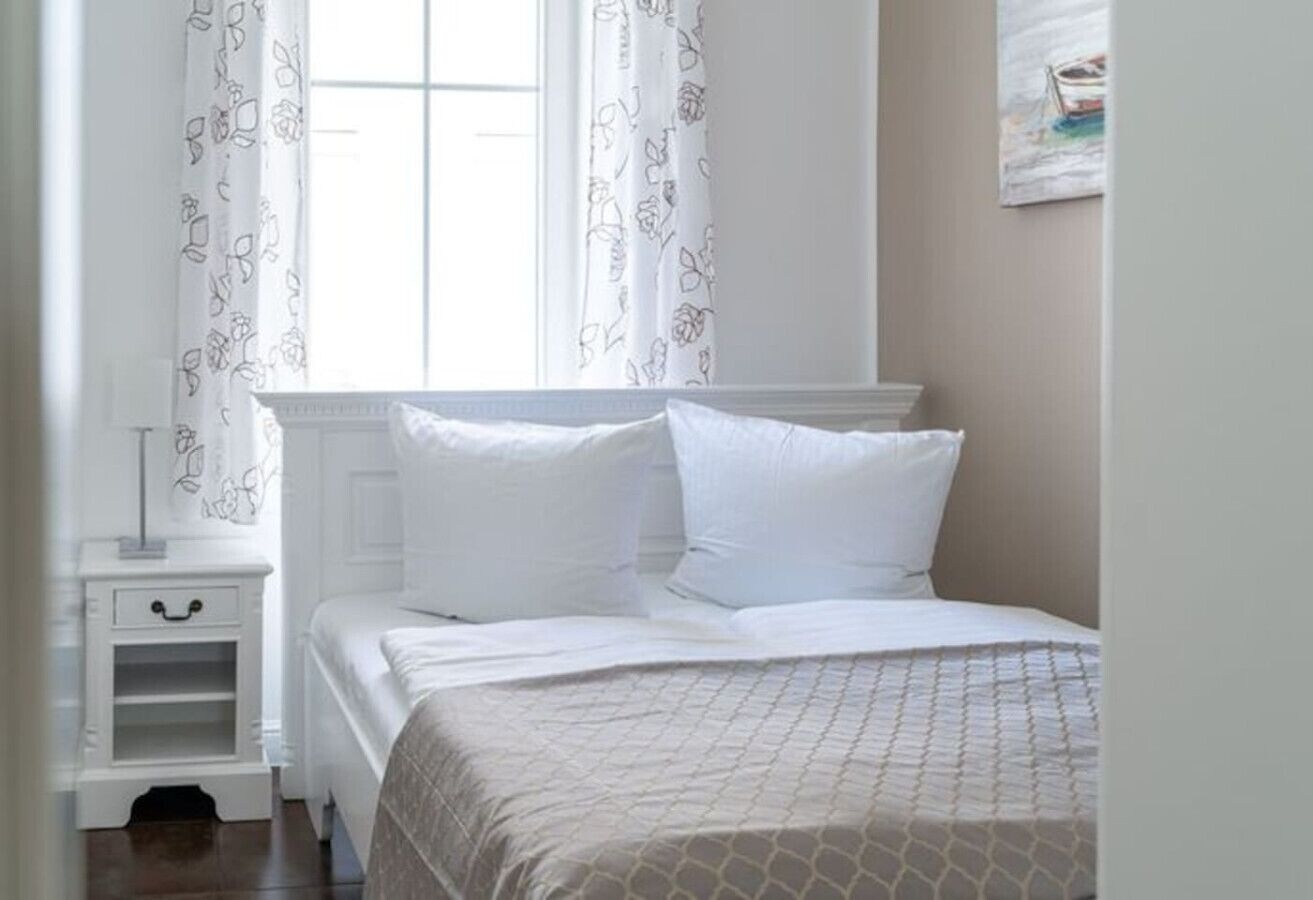Una cama bien hecha con ropa blanca y una colcha estampada, una mesita de noche con una lámpara, y una ventana con cortinas florales.