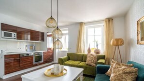 Una sala moderna con un sofá verde, una mesa de centro blanca y una cocina con armarios de madera y un microondas.