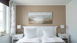 A bedroom with a bed, two bedside tables, a painting of a beach scene, and a window with a curtain. - Villa Meeresgold "Sea View" Premium Suite (Binz)