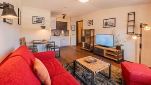 A modern living room with a red sofa, a coffee table, and a TV. There is a kitchen in the background with white cabinets and a dining area. - Inselzeit Comfort Suite No. 20 (Sellin)