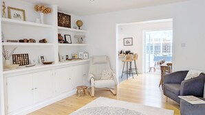 A spacious living room with a white sofa, a grey armchair, and a coffee table. There are shelves with various decorative items and a dining area visible through an open door. - A family house that sleeps 6 guests  in 4 bedrooms (Chichester)
