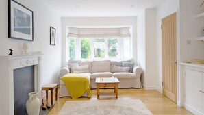 A bright living room with a fireplace, a sofa, a small table, and a view through a bay window. - A family house that sleeps 6 guests  in 4 bedrooms (Chichester)