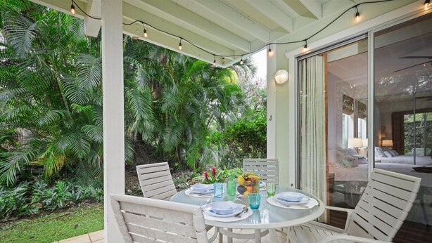 Une terrasse couverte avec une table ronde dressée pour le repas, entourée de chaises blanches, et une porte vitrée donnant sur une chambre.