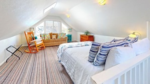 A bedroom with a sloped ceiling, a bed with striped pillows, a rocking chair, a small table with a lamp, and a window with blinds.