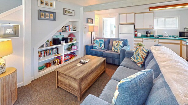 A cozy living room with a sofa, coffee table, and bookshelves. A kitchen with a refrigerator and stove is visible in the background.
