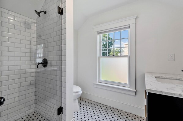 Une salle de bain avec des carreaux de céramique blancs rectangulaires, une pomme de douche noire, des toilettes et une fenêtre donnant sur des arbres.