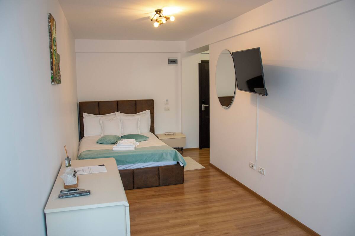 Una habitación de hotel moderna con piso de madera, una cama con cubrecama verde, una mesita de noche con control remoto y un televisor de pantalla plana montado en la pared.