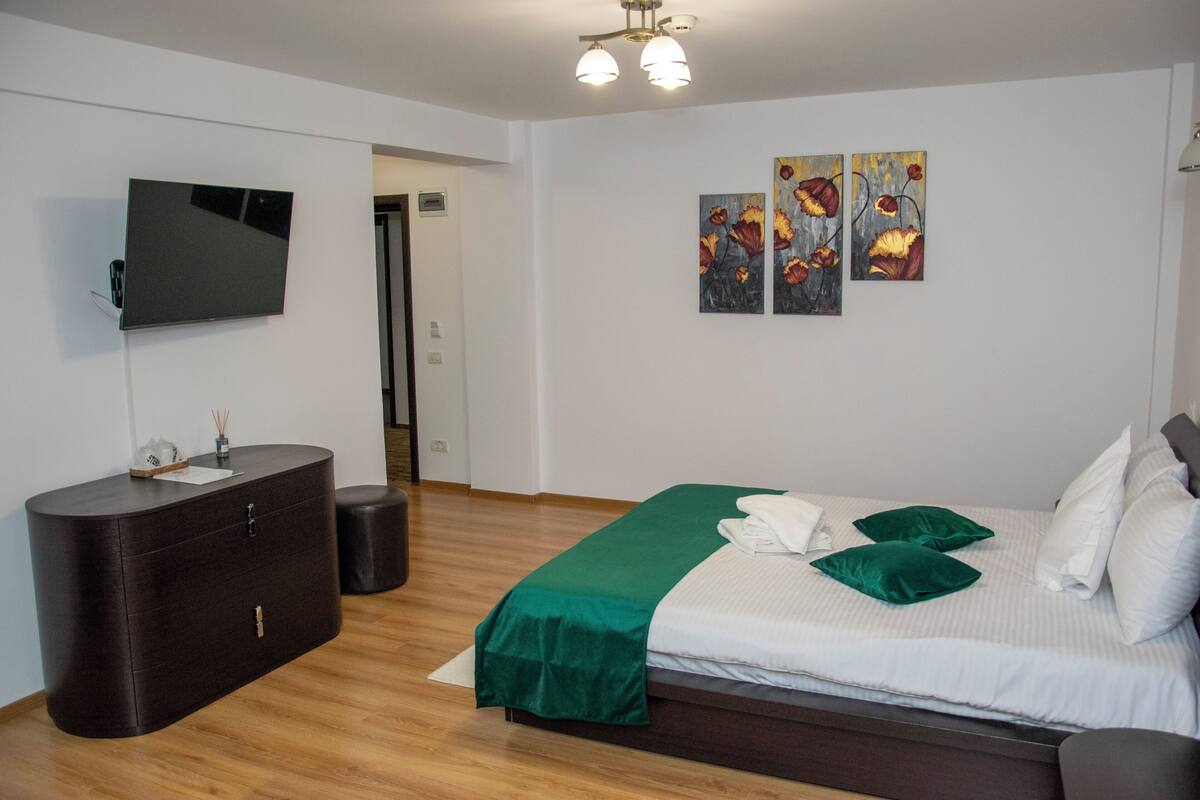 Habitación de hotel moderna con televisión de pantalla plana, cómoda de madera, cama con ropa de cama verde y dos cuadros en la pared.