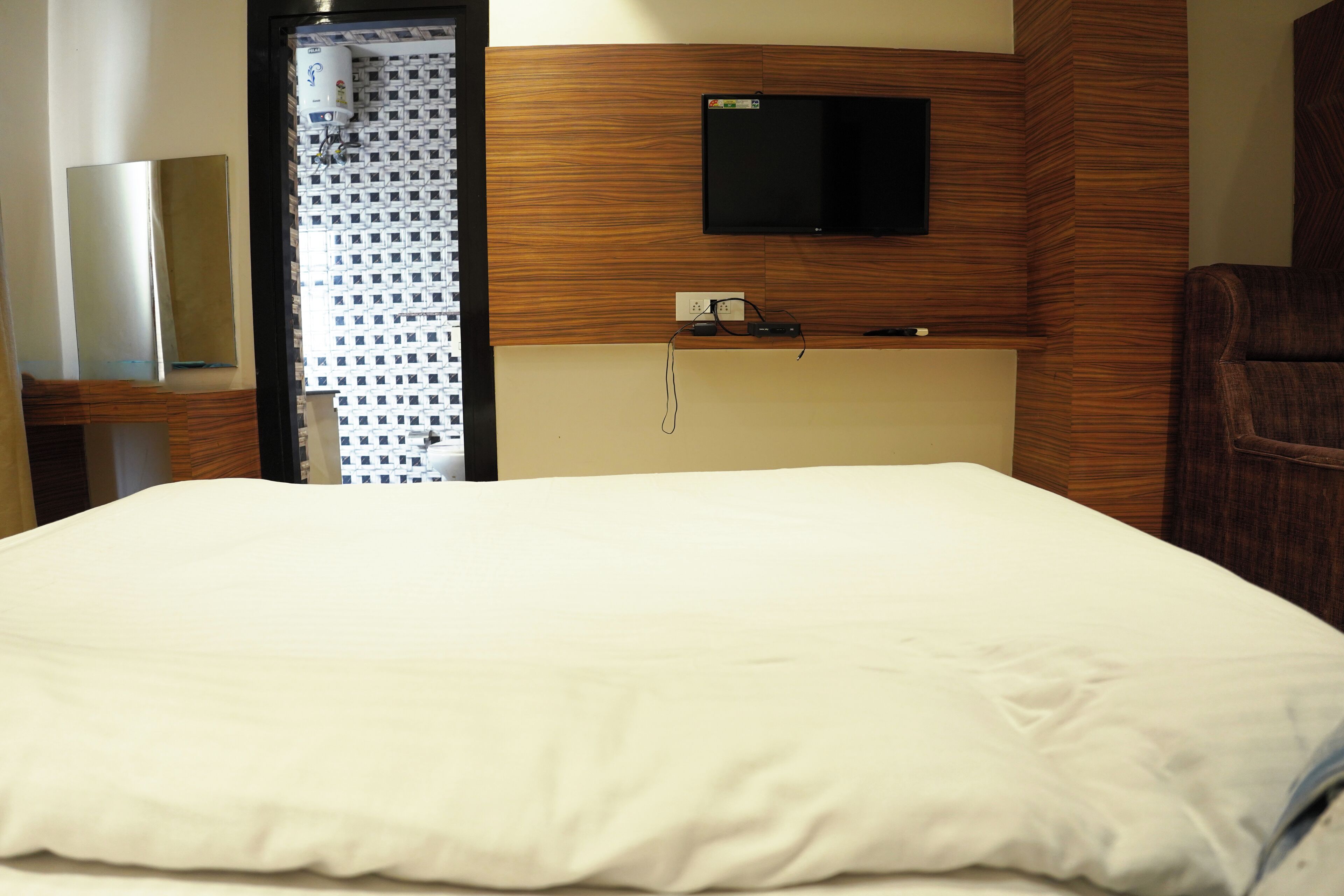 A hotel room with a bed, a flat-screen TV mounted on the wall, a wooden headboard, and a small table with a lamp.