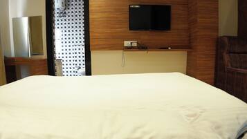 A hotel room with a bed, a flat-screen TV mounted on the wall, a wooden headboard, and a small table with a lamp.