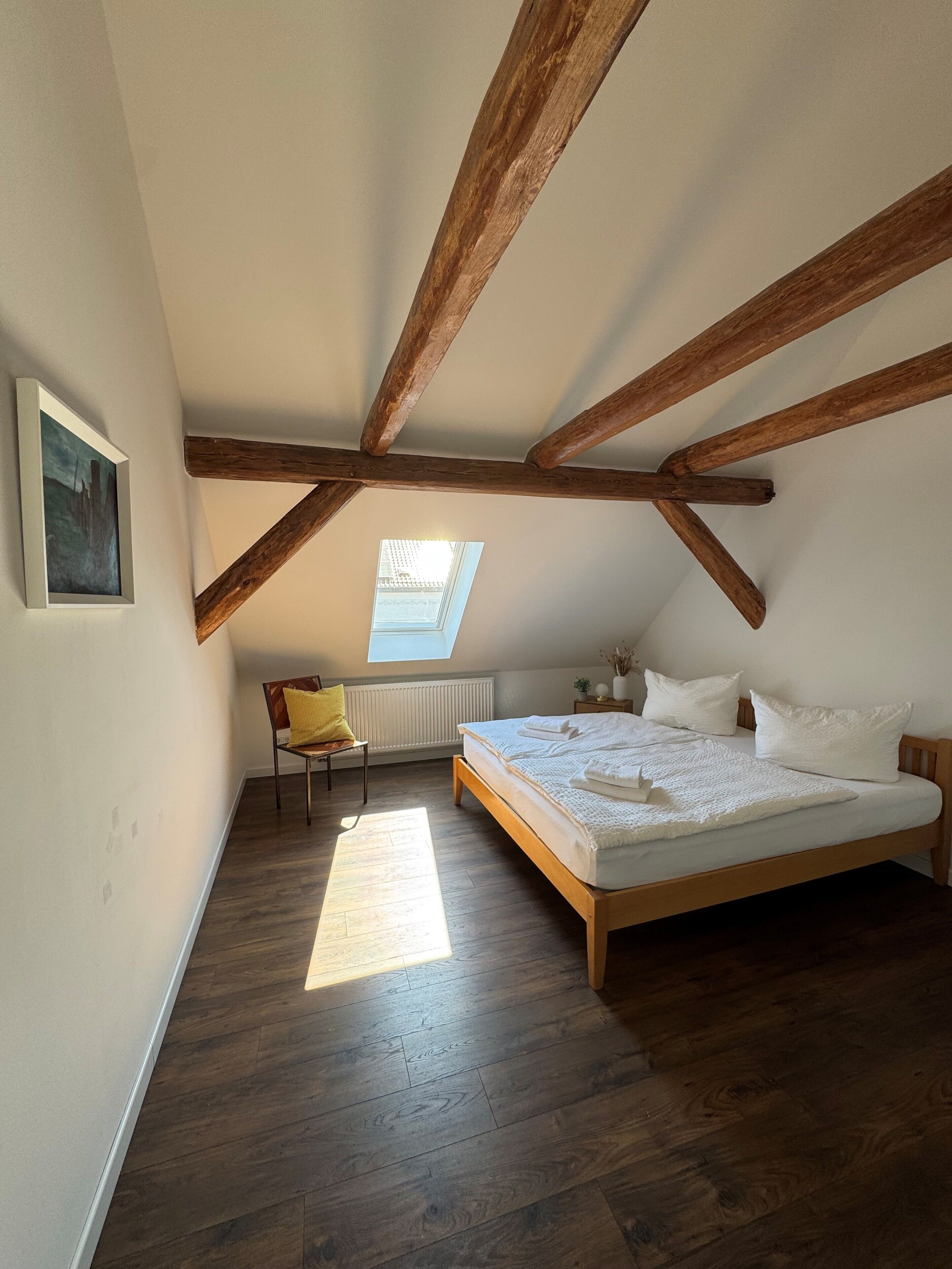 Ein Schlafzimmer mit schrägem Dach, Holzbalken, einem Oberlicht, einem Bett, einem Sessel und einer Heizung.