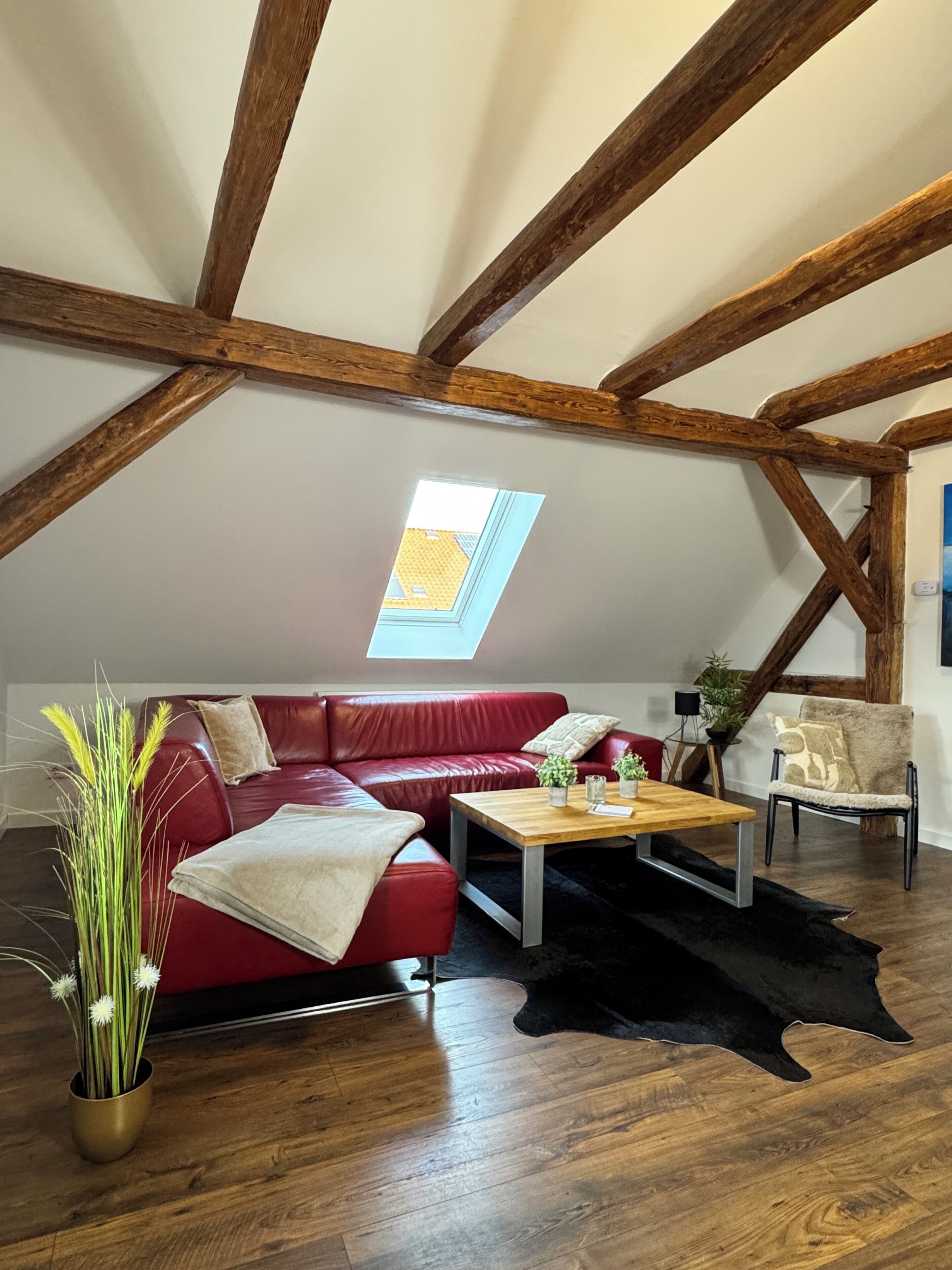 Ein Dachzimmer mit freiliegenden Holzbalken, einem roten Ledersofa, einem Holztisch und einem schwarzen Teppich sowie einem Oberlicht.