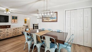 Zona pranzo con tavolo in legno, sedie azzurre e un lampadario. Si nota un divano e una televisione sullo sfondo.