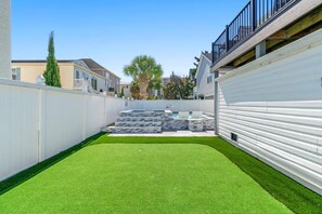 Patio trasero residencial con césped artificial, cerca blanca y una escalera que lleva a una terraza.