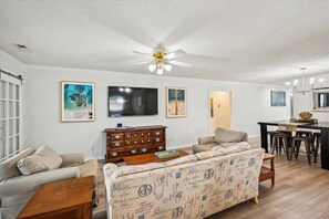 Una sala de estar con un sofá, televisor, ventilador de techo y comedor con mesa y sillas.