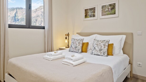 Una cama bien hecha con sábanas blancas y almohadas estampadas con motivos florales, una mesita de noche con lámpara, y una ventana con vistas a la montaña.