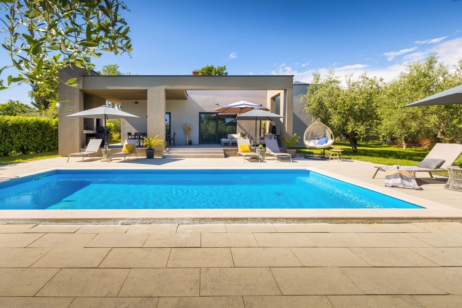 A modern pool area with a clear blue pool, lounge chairs, umbrellas, and a covered seating area.