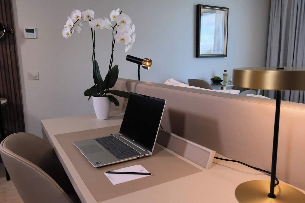 Una habitación de hotel moderna con un ordenador portátil sobre un escritorio, una orquídea en maceta, una lámpara de pie y un cuadro en la pared.