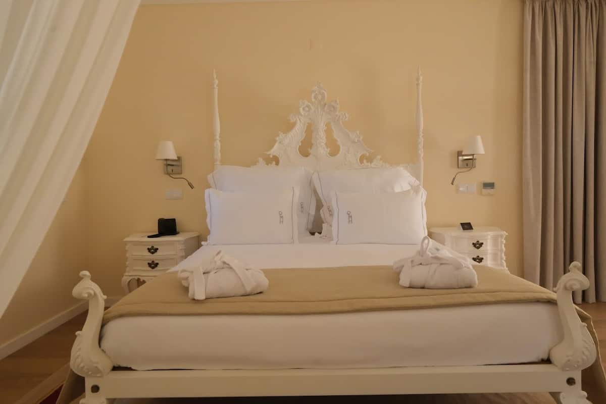 Una cama bien hecha con ropa blanca y dos toallas, un cabecero blanco ornamentado, dos mesitas de noche con lámparas y una pared color beige.