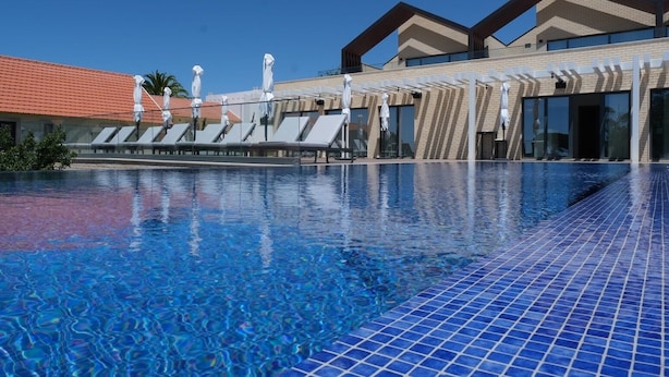 Uma piscina com espreguiçadeiras e guarda-sóis, um deck revestido de azulejos, e um edifício com telhado de telhas vermelhas e grandes janelas.