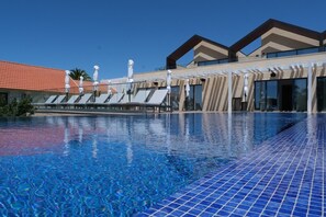 Une piscine avec des chaises longues et des parasols, une terrasse carrelée et un bâtiment doté d’un toit recouvert de tuiles rouges et de grandes fenêtres.