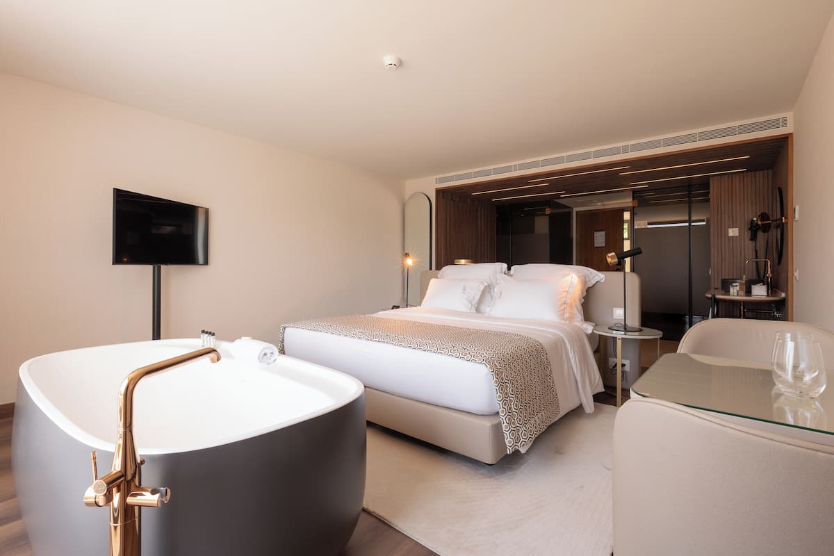 Una habitación de hotel moderna con una bañera grande, una cama con cubrecama estampado, una mesita de cristal y un televisor montado en la pared.
