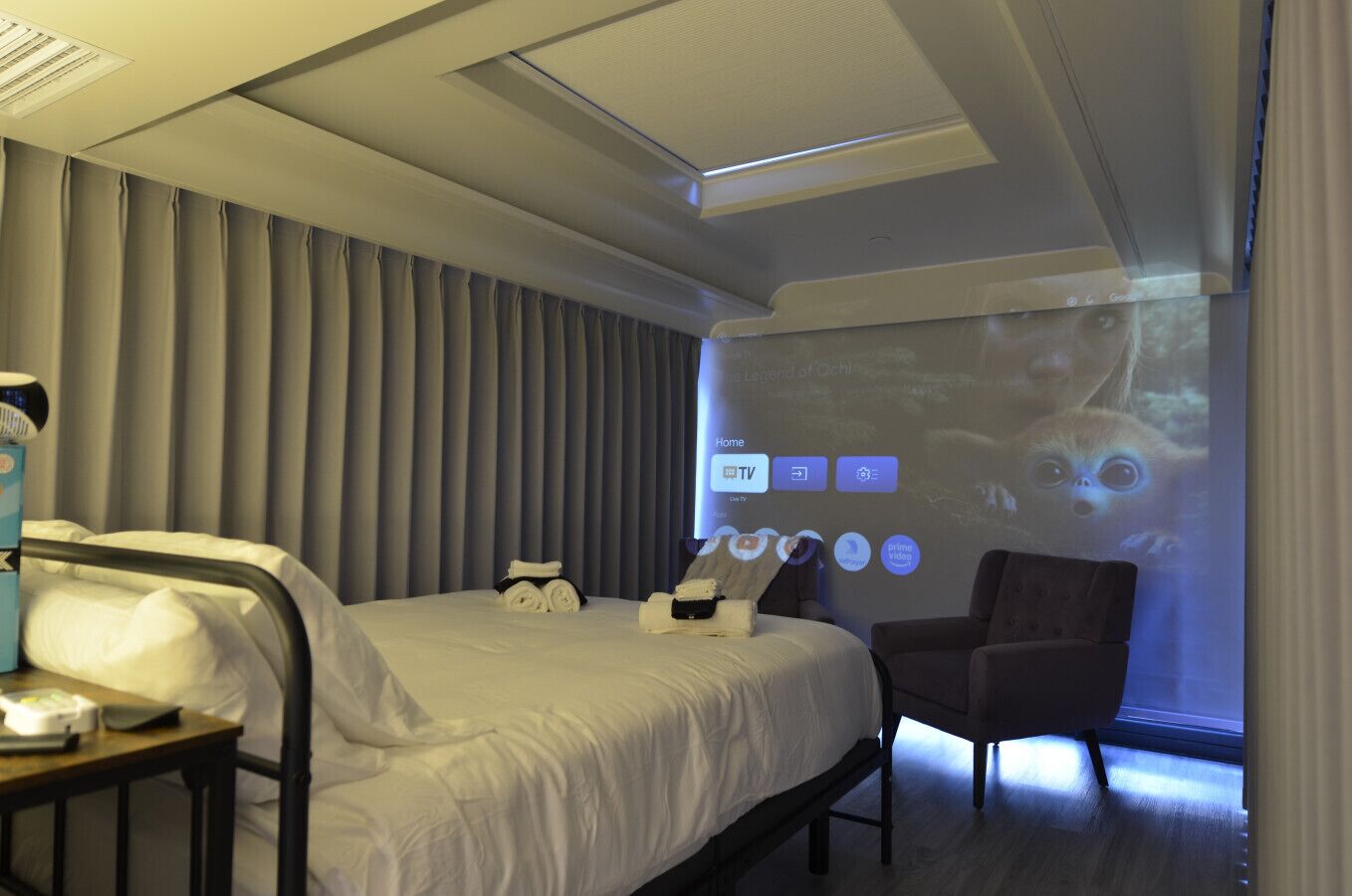 A hotel room with a bed, a desk, a chair, and a large screen displaying a TV interface.