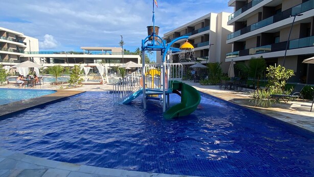 Une piscine avec un toboggan aquatique et un toboggan vert, entourée d’une terrasse carrelée et d’un immeuble à plusieurs étages doté de balcons.
