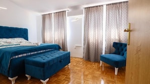 A bedroom with a large bed, two blue upholstered chairs, a wooden floor, and curtains.