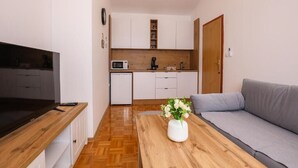 A compact living space with a kitchenette, a flat-screen TV, a grey sofa, and a wooden coffee table with a vase of flowers.