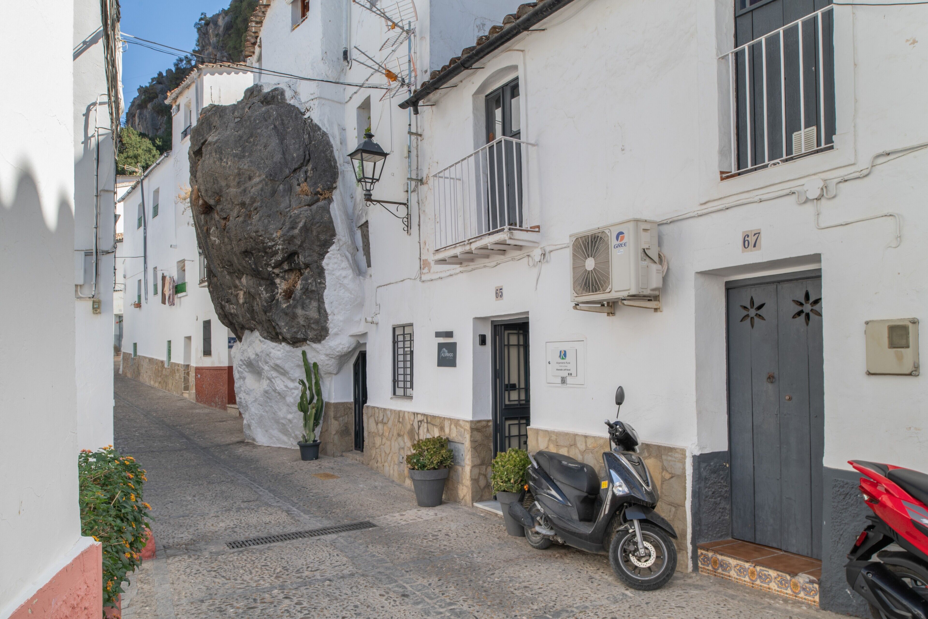 A narrow street with cobblestone pavement, white buildings, a large rock formation, and a parked scooter.