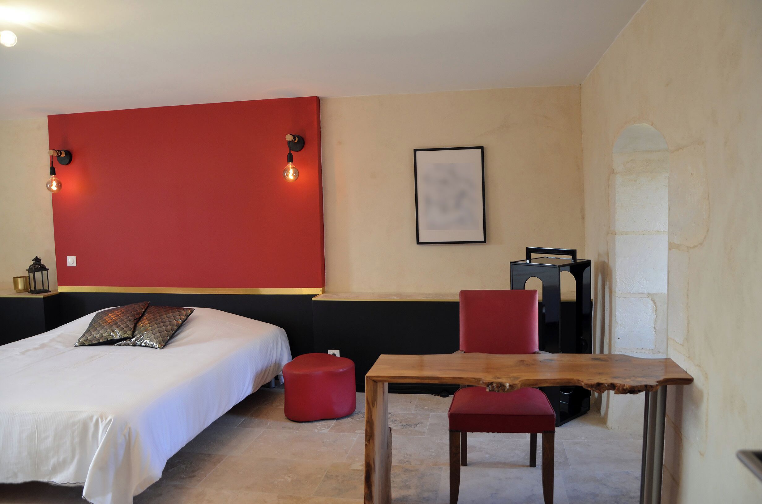 A hotel room with a bed, a wooden table, a red ottoman, and a black cat on the table.