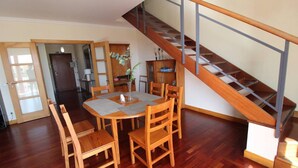 Un comedor con una mesa y sillas de madera, una escalera con barandilla de metal y vista a la cocina en el fondo.