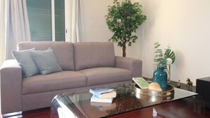 Un salón con un sofá color beige, una mesa de centro de cristal con libros y objetos decorativos, una planta en maceta y una ventana con persianas.