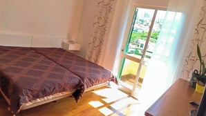 A bedroom with a bed, a bedside table, a window with curtains, and a view of the outside. - Jardim Sol Holiday Apartment, Jardim Sol Holiday Apartment (Funchal)