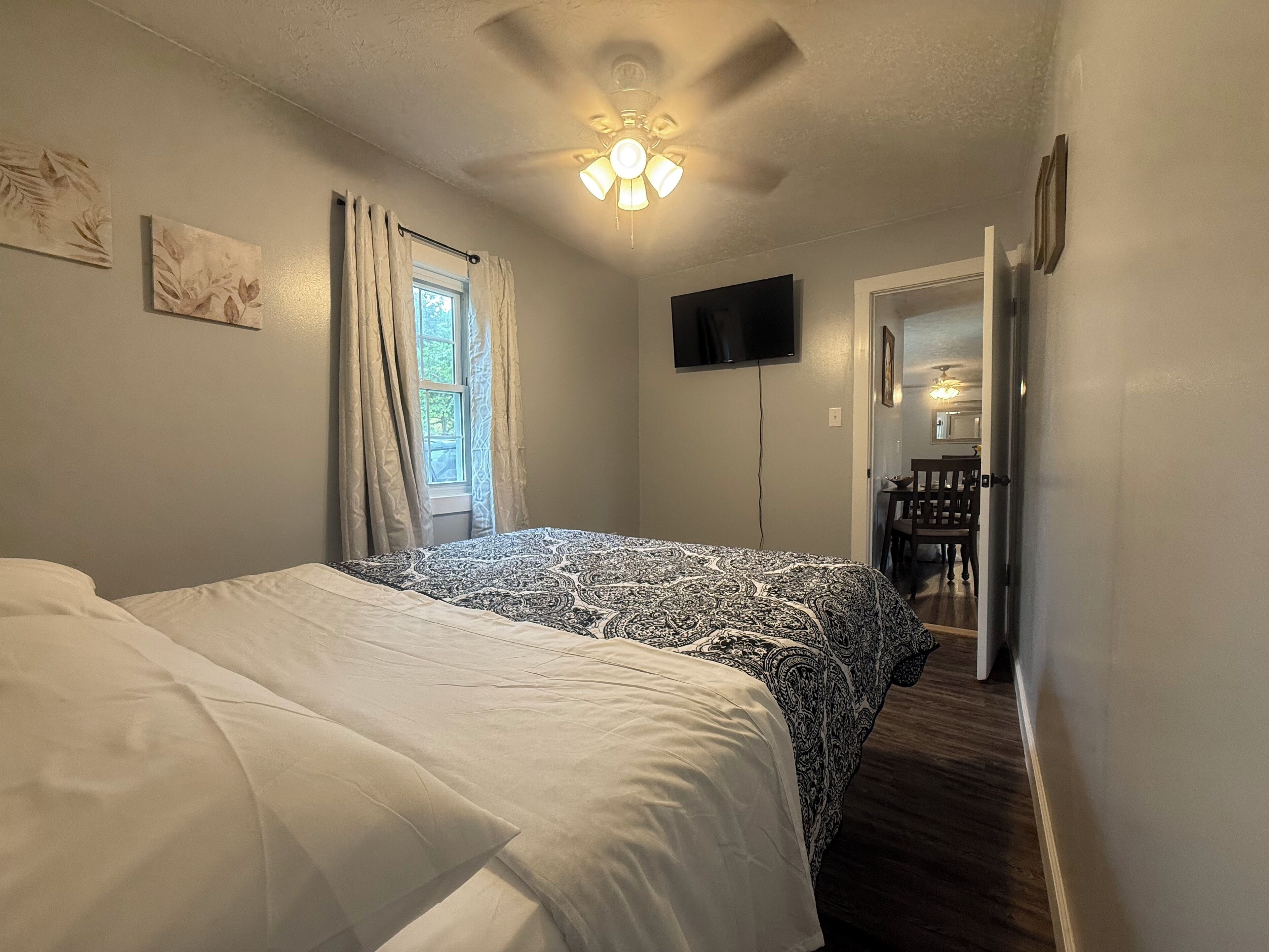 A bedroom with a bed, a ceiling fan, a TV, and a door leading to a dining area.