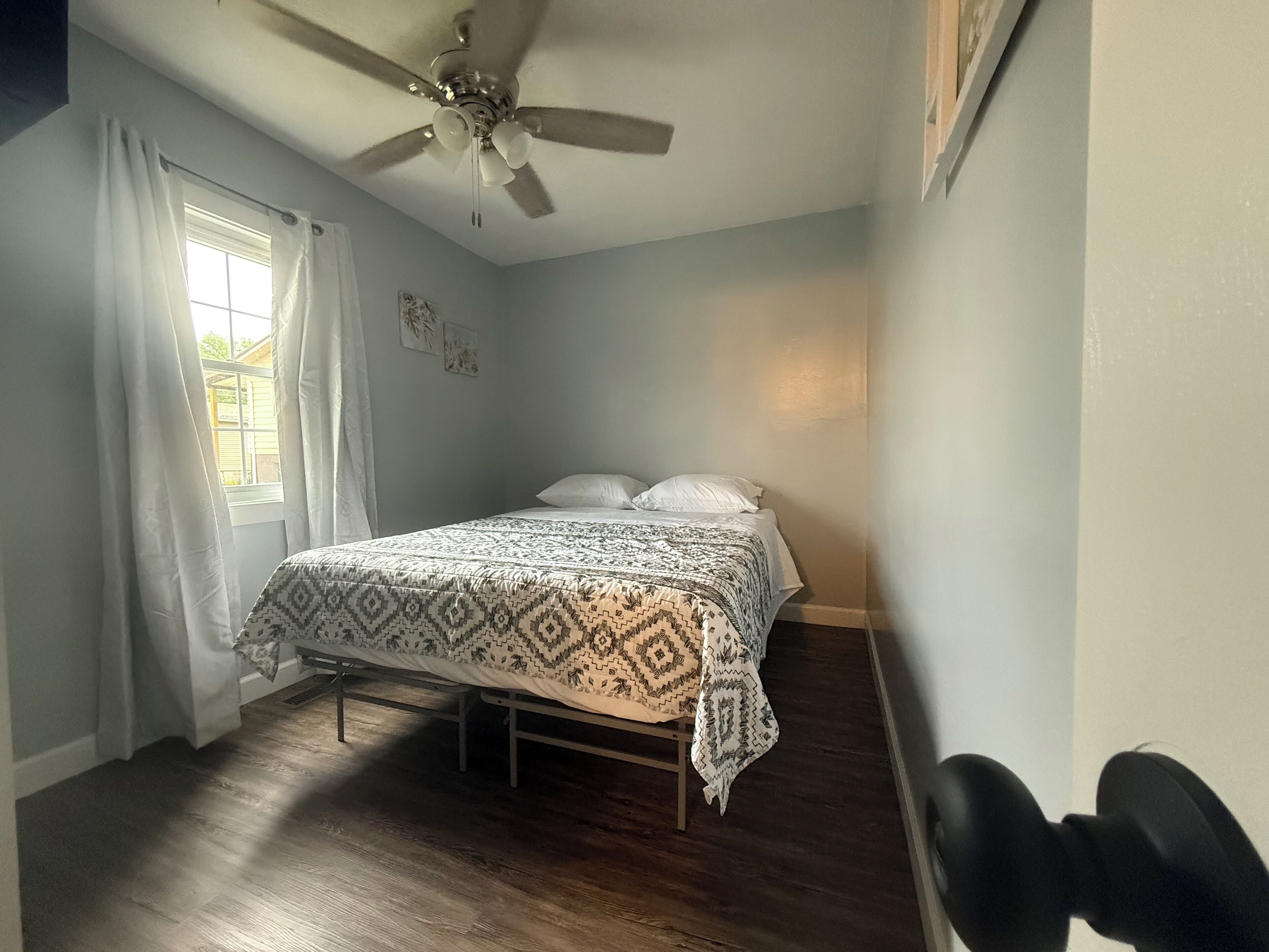 Une chambre à coucher avec un lit, des rideaux blancs, un ventilateur de plafond et une fenêtre.