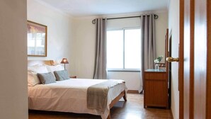A bedroom with a bed, bedside lamp, window with curtains, and a wooden dresser.