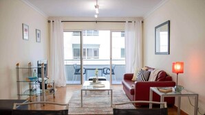 A modern living room with a glass coffee table, a red sofa, a glass TV stand, and a balcony with a view.