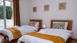 A hotel room with two beds, each with white bedding and a mustard-colored throw blanket. There are two bedside tables with lamps, and the room has a large window with curtains.