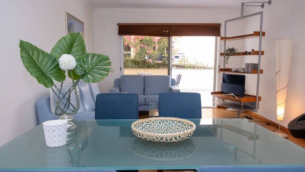 A modern living room with a glass-top table, blue chairs, a sofa, and a TV on a wooden shelf.