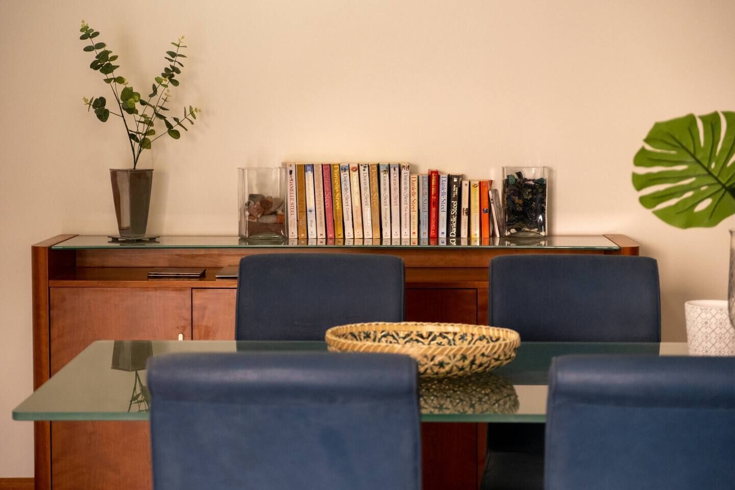 A dining area with a glass table, blue chairs, a bookshelf with books, a potted plant, and a decorative bowl.