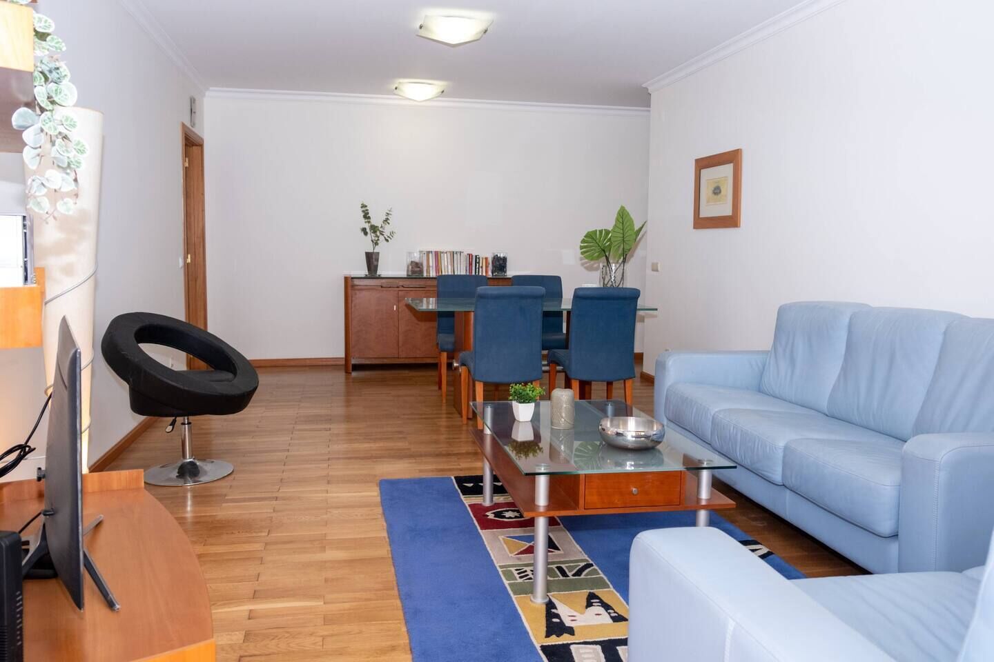 Un salón con un sofá azul, una mesa de centro de vidrio, una mesa de comedor con sillas, una planta pequeña y un taburete anular.