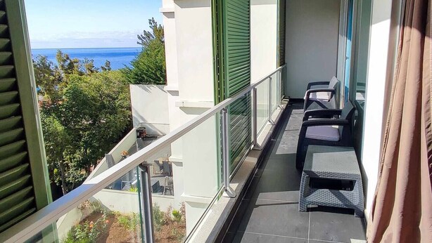 Un balcón con vista al mar, barandilla de cristal y mobiliario exterior.