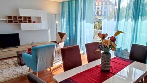 Una sala de estar moderna con televisor, una mesa de comedor con sillas, un jarrón de flores y vistas a edificios a través de amplios ventanales.